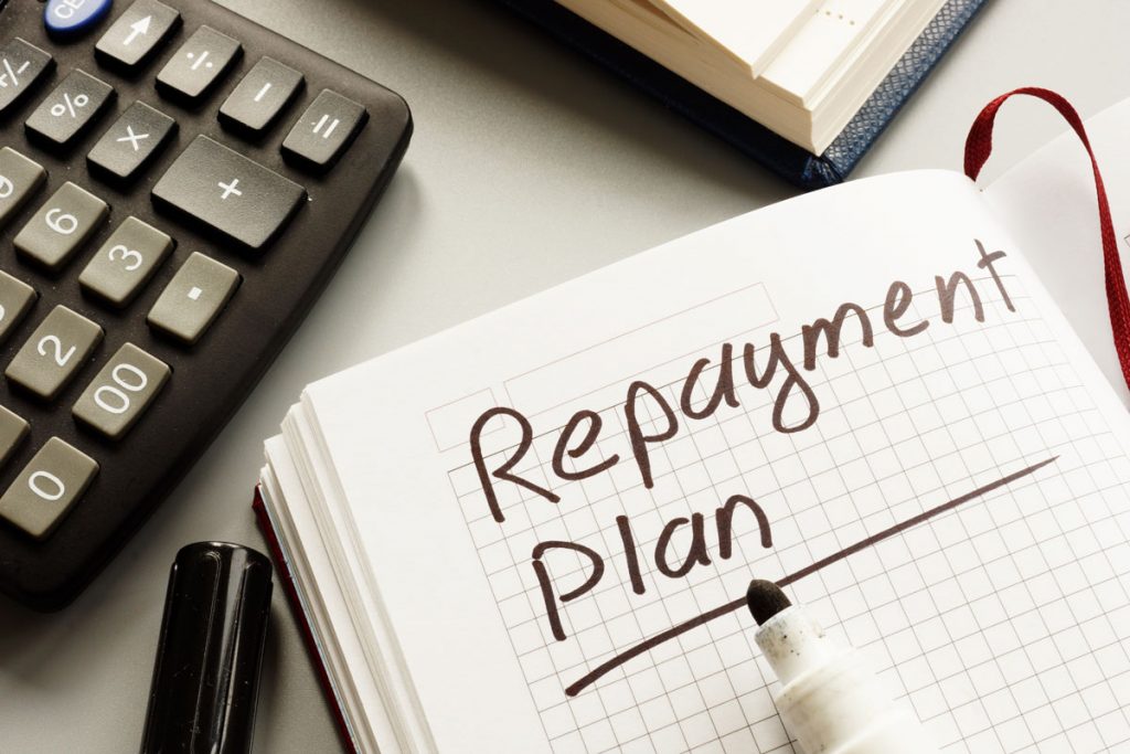 The words Repayment Plan written on a notebook, and calculator on desk beside it as lowest interest rates are being searched for